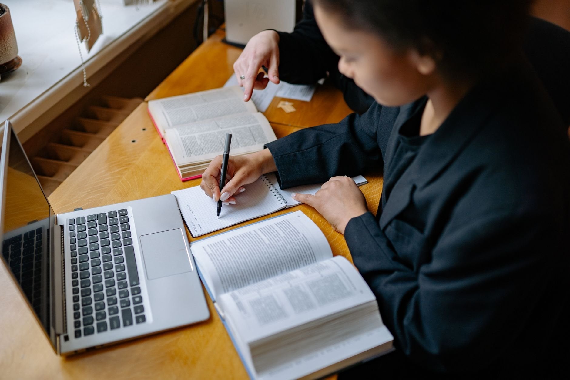 Two students engaged in study, taking notes with books and a laptop on a wooden desk indoors.